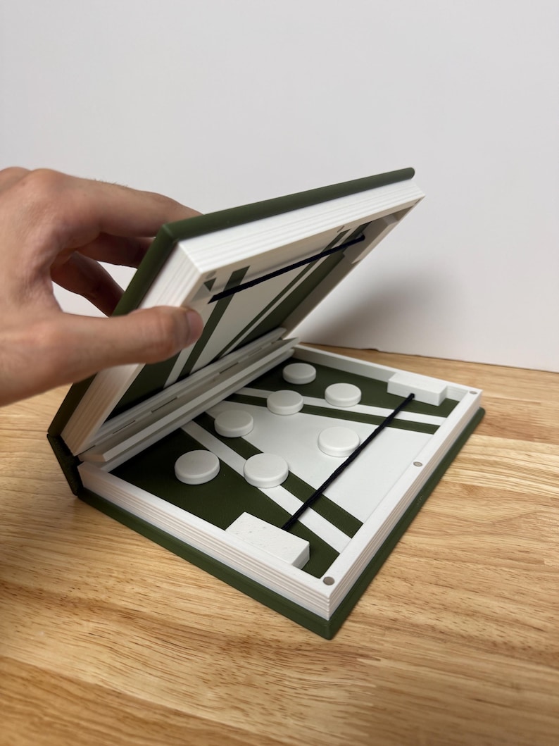 Peut inclure: Un jeu de soci&eacute;t&eacute; vert et blanc dans un &eacute;tui en forme de livre. Le plateau de jeu pr&eacute;sente un motif vert et blanc avec des lignes noires et des pions blancs. L'&eacute;tui est ouvert, r&eacute;v&eacute;lant le jeu.