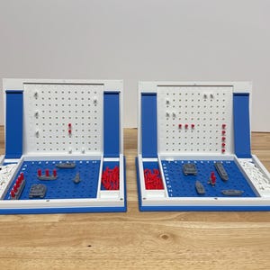 Peut inclure: Deux jeux de soci&eacute;t&eacute; Battleship c&ocirc;te &agrave; c&ocirc;te. Chaque jeu a un champ de jeu bleu avec des navires miniatures et des chevilles rouges et blanches. Les plateaux de jeu sont blancs avec des accents bleus et sont ouverts.