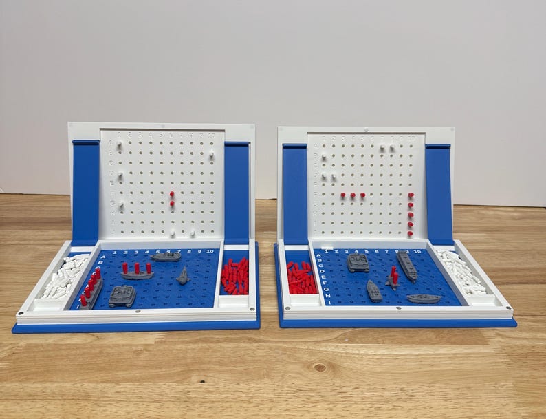 Peut inclure: Deux jeux de soci&eacute;t&eacute; Battleship c&ocirc;te &agrave; c&ocirc;te. Chaque jeu a un champ de jeu bleu avec des navires miniatures et des chevilles rouges et blanches. Les plateaux de jeu sont blancs avec des accents bleus et sont ouverts.
