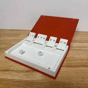 Peut inclure: Un jeu de Shut the Box rouge et blanc. Le jeu est ouvert, r&eacute;v&eacute;lant un int&eacute;rieur blanc avec des tuiles num&eacute;rot&eacute;es et deux d&eacute;s. Les tuiles sont num&eacute;rot&eacute;es de 1 &agrave; 12. Les d&eacute;s montrent les chiffres 2 et 5.