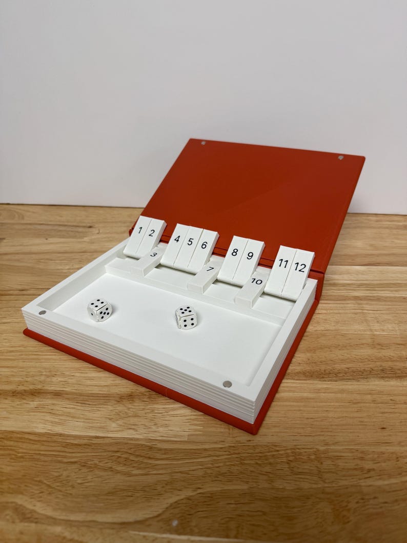 Peut inclure: Un jeu de Shut the Box rouge et blanc. Le jeu est ouvert, r&eacute;v&eacute;lant un int&eacute;rieur blanc avec des tuiles num&eacute;rot&eacute;es et deux d&eacute;s. Les tuiles sont num&eacute;rot&eacute;es de 1 &agrave; 12. Les d&eacute;s montrent les chiffres 2 et 5.