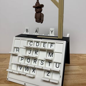 Peut inclure: Un jeu de mots sur le th&egrave;me du gibet. Le jeu comprend un tableau de lettres blanc avec des lettres noires, une figurine miniature pendue et une structure de potence en bois. Les lettres "B", "A" et "Y" sont affich&eacute;es au-dessus du tableau.