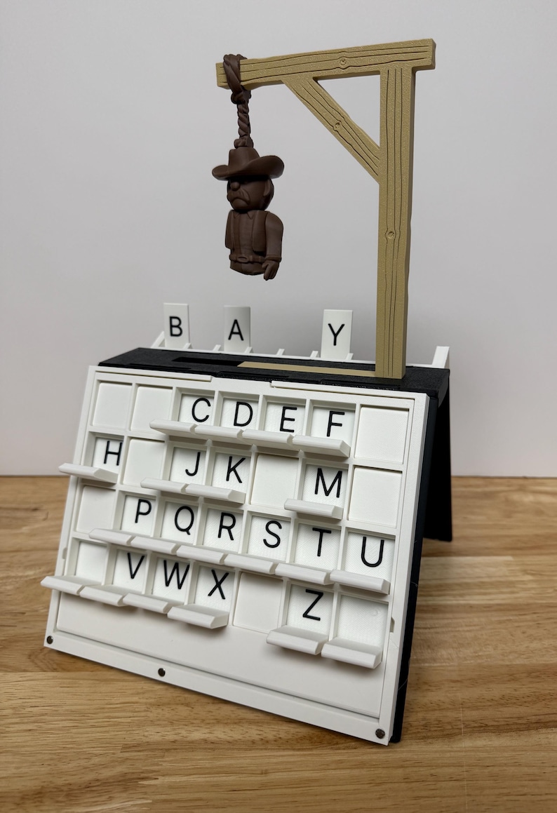 Peut inclure: Un jeu de mots sur le th&egrave;me du gibet. Le jeu comprend un tableau de lettres blanc avec des lettres noires, une figurine miniature pendue et une structure de potence en bois. Les lettres "B", "A" et "Y" sont affich&eacute;es au-dessus du tableau.