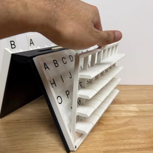 Peut inclure: Un organiseur d'alphabet blanc et noir avec une main ouvrant une section. L'organisateur comporte plusieurs &eacute;tag&egrave;res avec des lettres imprim&eacute;es dessus. Les lettres sont en noir et les &eacute;tag&egrave;res sont blanches. L'organisateur est sur une surface en bois.