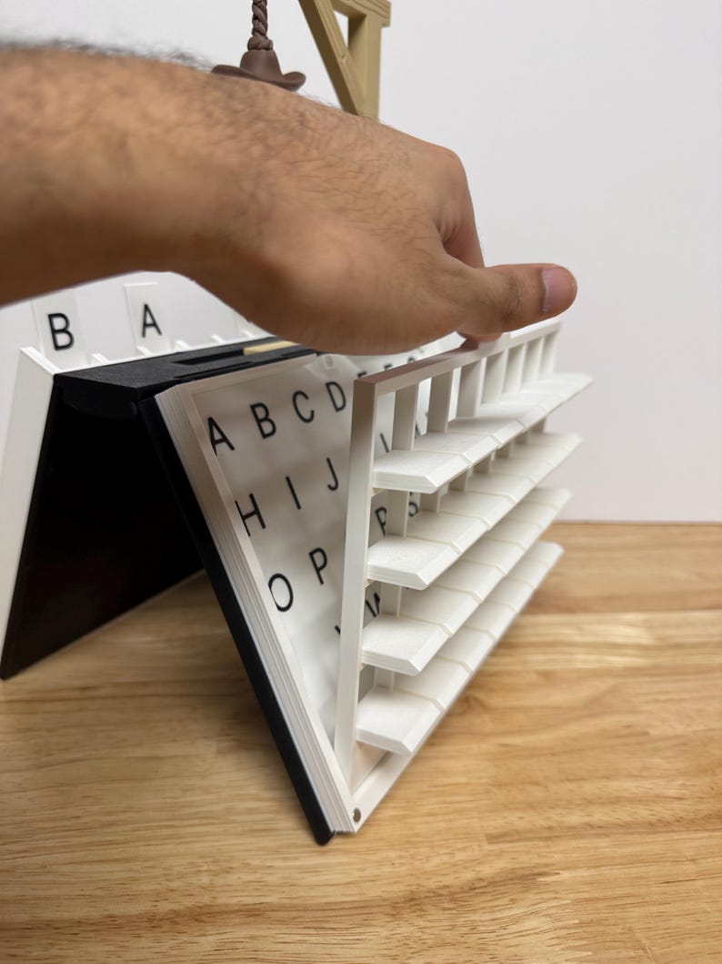 Peut inclure: Un organiseur d'alphabet blanc et noir avec une main ouvrant une section. L'organisateur comporte plusieurs &eacute;tag&egrave;res avec des lettres imprim&eacute;es dessus. Les lettres sont en noir et les &eacute;tag&egrave;res sont blanches. L'organisateur est sur une surface en bois.