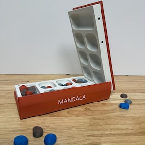 Peut inclure: Un jeu de soci&eacute;t&eacute; Mancala ouvert, avec des accents rouges et blancs. Le plateau de jeu comporte plusieurs compartiments remplis de pierres color&eacute;es. Le mot "MANCALA" est imprim&eacute; sur le c&ocirc;t&eacute; du plateau. Plusieurs pierres bleues et grises sont &eacute;parpill&eacute;es autour du plateau.