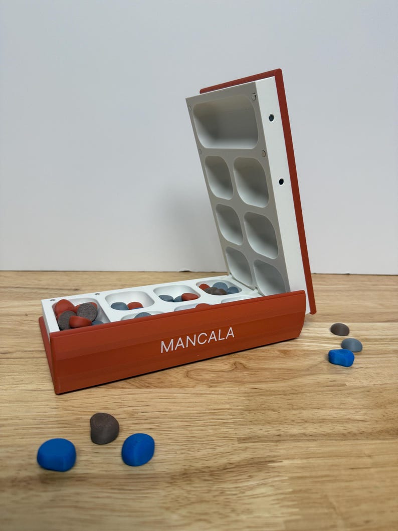 Peut inclure: Un jeu de soci&eacute;t&eacute; Mancala ouvert, avec des accents rouges et blancs. Le plateau de jeu comporte plusieurs compartiments remplis de pierres color&eacute;es. Le mot "MANCALA" est imprim&eacute; sur le c&ocirc;t&eacute; du plateau. Plusieurs pierres bleues et grises sont &eacute;parpill&eacute;es autour du plateau.