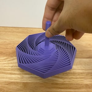 May include: A purple, geometric-patterned spinning top. The top has a hexagonal base with layered, angled lines that create a spiral effect. The top is held by a hand, showing the central peg.