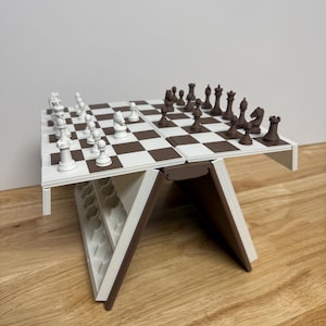 Peut inclure: Un jeu d'&eacute;checs pliable blanc et marron, avec des pi&egrave;ces dispos&eacute;es sur l'&eacute;chiquier. Le plateau est blanc avec des cases marron. Les pi&egrave;ces sont blanches et marron. L'ensemble est pos&eacute; sur une surface en bois.