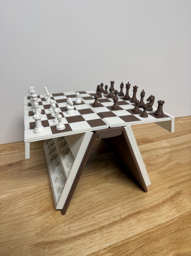 Peut inclure: Un jeu d'&eacute;checs pliable blanc et marron, avec des pi&egrave;ces dispos&eacute;es sur l'&eacute;chiquier. Le plateau est blanc avec des cases marron. Les pi&egrave;ces sont blanches et marron. L'ensemble est pos&eacute; sur une surface en bois.