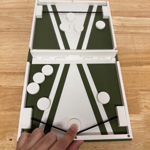 Peut inclure: Jeu de soci&eacute;t&eacute; en bois avec un design vert et blanc. Le jeu est ouvert, r&eacute;v&eacute;lant une surface de jeu avec des pions circulaires blancs. Une main touche l'un des pions. Le jeu est sur une surface en bois.