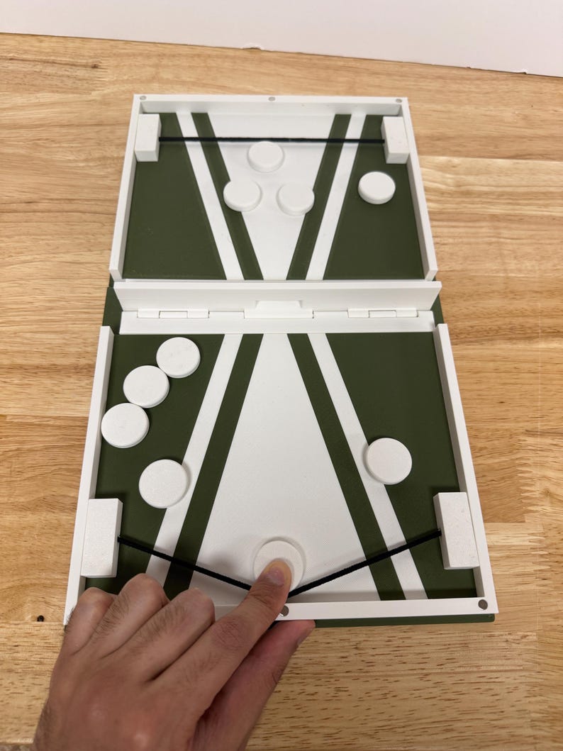 Peut inclure: Jeu de soci&eacute;t&eacute; en bois avec un design vert et blanc. Le jeu est ouvert, r&eacute;v&eacute;lant une surface de jeu avec des pions circulaires blancs. Une main touche l'un des pions. Le jeu est sur une surface en bois.
