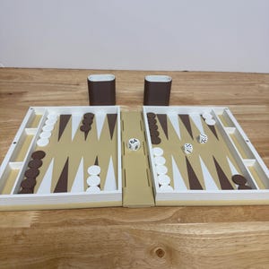 Peut inclure: Un jeu de backgammon avec une surface de jeu beige, des pions blancs et marron, et deux gobelets &agrave; d&eacute;s marron. Les d&eacute;s affichent les chiffres 6, 4, 8, 2 et 3. Le plateau de jeu est ouvert et pr&ecirc;t &agrave; jouer.