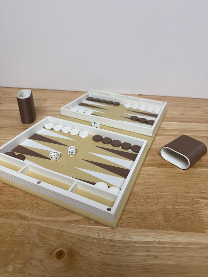 Peut inclure: Un jeu de backgammon avec une surface de jeu beige, des marquages triangulaires marron et blancs, et des pions blancs et marron. Deux d&eacute;s sont sur le plateau. Deux gobelets &agrave; d&eacute;s marron sont &eacute;galement pr&eacute;sents. L'ensemble est pos&eacute; sur une surface en bois.