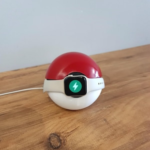 May include: A white and red Pokeball-shaped charging stand for an Apple Watch. The watch is charging and the screen shows a green lightning bolt icon.