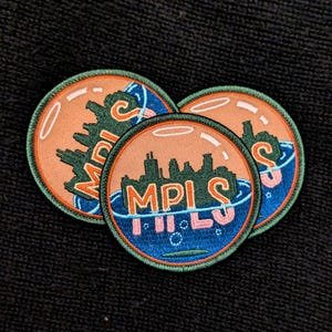 May include: Three circular embroidered patches. Each patch features a cityscape silhouette in green, the word "MPLS" in pink and orange, and a blue water design. The patches have an orange background and a green border.