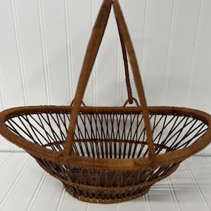 May include: A brown wicker basket with a handle. The basket has an oval shape and is made of woven wicker. The handle is attached to the sides of the basket and is also made of woven wicker. The basket is empty.