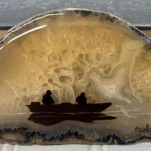 May include: A polished agate slice with a painted scene of two people in a boat on water. The agate features shades of tan, brown, and white, with a dark border. The boat and figures are silhouetted in black.