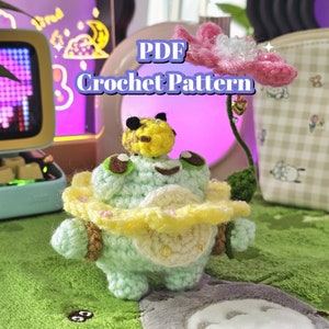 May include: Crochet pattern PDF for a green and yellow frog amigurumi with a bee on its head, holding a pink flower. The frog is wearing a yellow and white crocheted skirt.