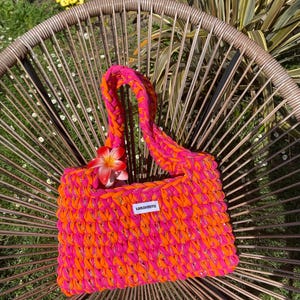May include: A rectangular crochet handbag crafted from vibrant neon pink and bright orange yarn. The bag features a thick braided handle and a small white rectangular label with the black text "IONAKNITS" attached to the front center.