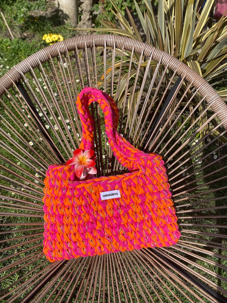 May include: A rectangular crochet handbag crafted from vibrant neon pink and bright orange yarn. The bag features a thick braided handle and a small white rectangular label with the black text "IONAKNITS" attached to the front center.