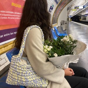 May include: A handmade crochet shoulder bag featuring a thick, braided strap and a woven pattern in alternating pastel yellow and light purple yarn. The bag includes a small rectangular white fabric label with black text on the front exterior.