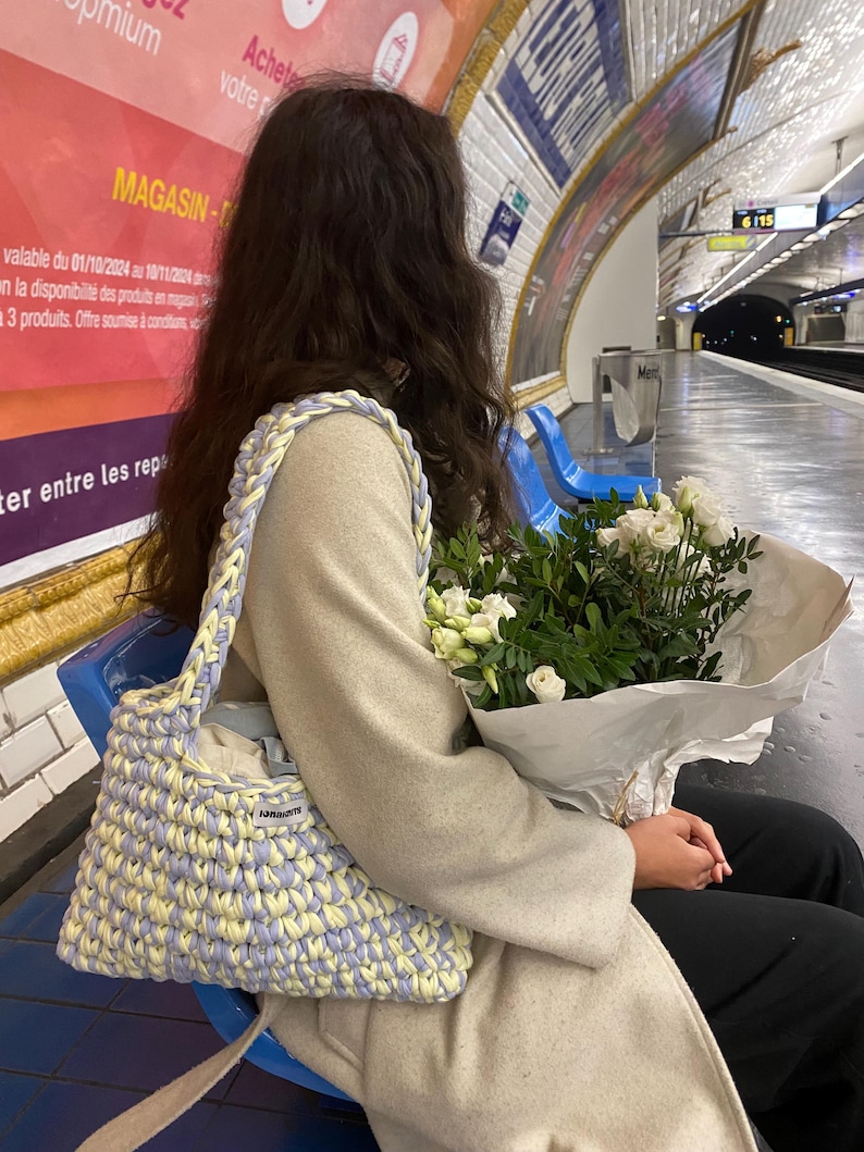 May include: A handmade crochet shoulder bag featuring a thick, braided strap and a woven pattern in alternating pastel yellow and light purple yarn. The bag includes a small rectangular white fabric label with black text on the front exterior.
