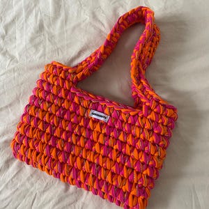 May include: A rectangular crochet handbag crafted from thick, vibrant neon orange and hot pink yarn interwoven with a thin metallic gold thread. The bag features a single integrated shoulder strap and a small white rectangular label with black text.