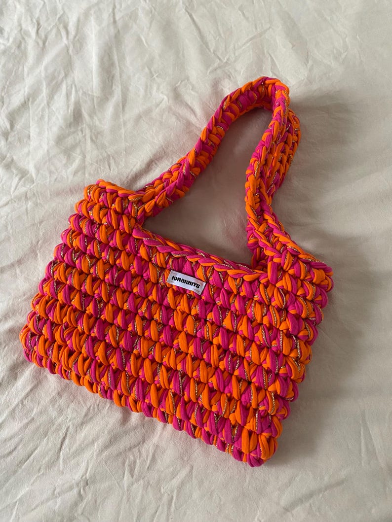May include: A rectangular crochet handbag crafted from thick, vibrant neon orange and hot pink yarn interwoven with a thin metallic gold thread. The bag features a single integrated shoulder strap and a small white rectangular label with black text.