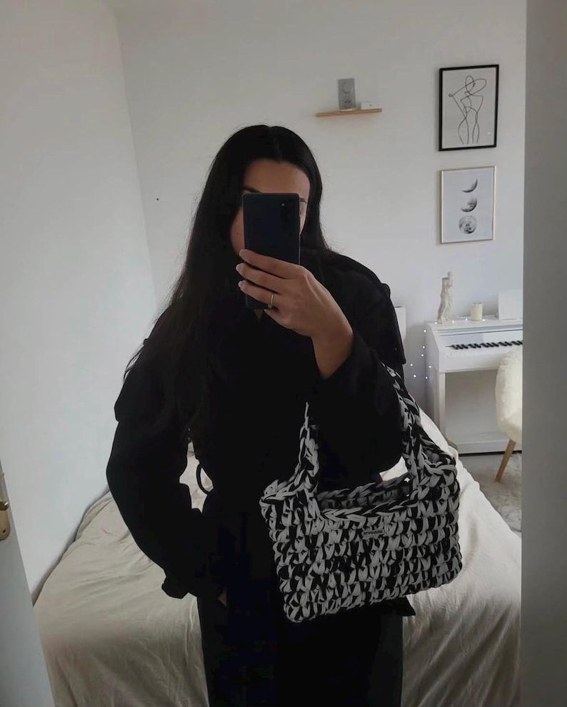 May include: A rectangular handbag featuring a thick, chunky knit texture with a high-contrast black and white marbled pattern. The bag includes two integrated top handles and a small rectangular brand label attached to the front surface.