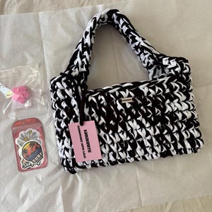 May include: A black and white crocheted handbag with a thick braided handle and a small rectangular metal logo plate. A pink tag attached to the handle reads "IONAKNITS HANDMADE WITH LOVE". Included are two small plastic bags containing pink flower charms and stickers.