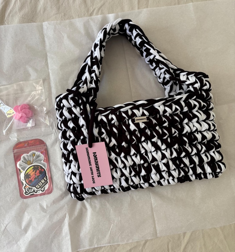 May include: A black and white crocheted handbag with a thick braided handle and a small rectangular metal logo plate. A pink tag attached to the handle reads "IONAKNITS HANDMADE WITH LOVE". Included are two small plastic bags containing pink flower charms and stickers.