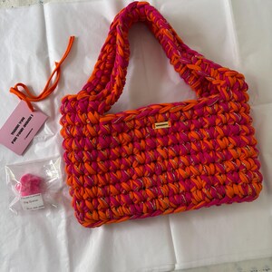 May include: A rectangular crochet handbag crafted from vibrant hot pink and bright orange yarn interwoven with a thin metallic gold thread. The bag features a single thick handle and a small rectangular gold-toned metal brand tag on the front.