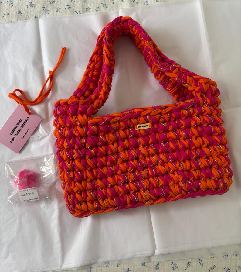 May include: A rectangular crochet handbag crafted from vibrant hot pink and bright orange yarn interwoven with a thin metallic gold thread. The bag features a single thick handle and a small rectangular gold-toned metal brand tag on the front.