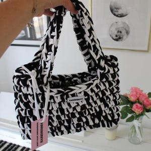 May include: A rectangular handbag crafted from thick black and white yarn in a chunky crochet stitch. The bag features two integrated handles and a small white fabric label reading "ionaknits". A pink rectangular tag is attached with white cord.