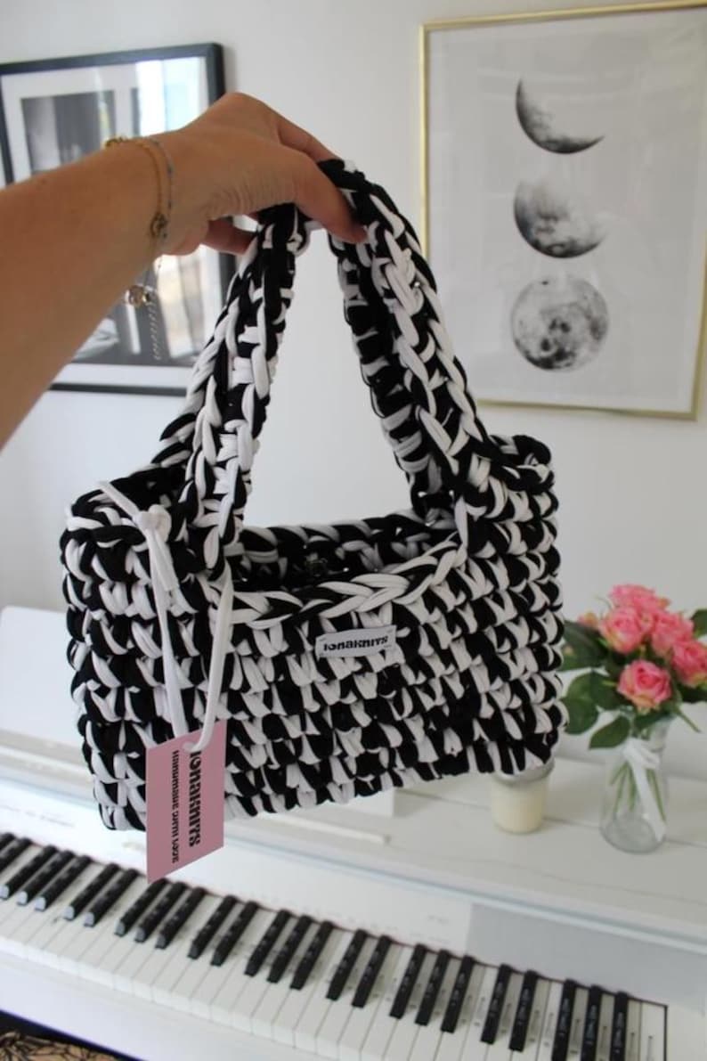 May include: A rectangular handbag crafted from thick black and white yarn in a chunky crochet stitch. The bag features two integrated handles and a small white fabric label reading "ionaknits". A pink rectangular tag is attached with white cord.