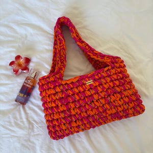 May include: A rectangular crochet handbag crafted from thick, vibrant pink and orange yarn interwoven with a subtle metallic gold thread. The bag features a single shoulder strap and a small rectangular gold-toned metal brand tag on the front.