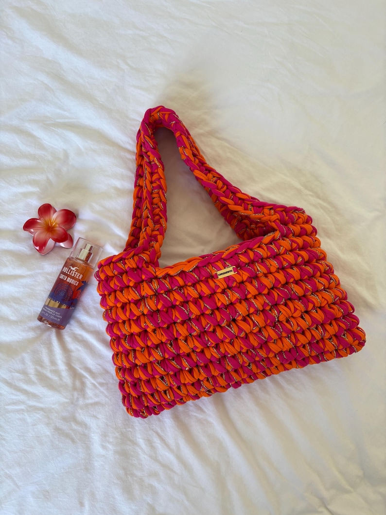 May include: A rectangular crochet handbag crafted from thick, vibrant pink and orange yarn interwoven with a subtle metallic gold thread. The bag features a single shoulder strap and a small rectangular gold-toned metal brand tag on the front.