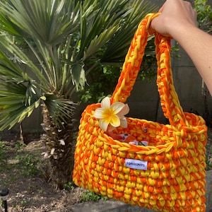 May include: A hand-crocheted tote bag made from thick, vibrant orange and yellow yarn. The bag features a single shoulder strap, a white fabric label with the text "IONAKNITS" in black, and a decorative white and yellow plumeria flower attached.