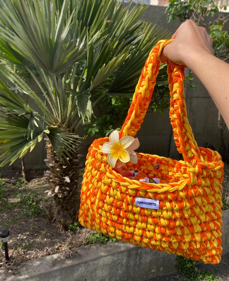 May include: A hand-crocheted tote bag made from thick, vibrant orange and yellow yarn. The bag features a single shoulder strap, a white fabric label with the text "IONAKNITS" in black, and a decorative white and yellow plumeria flower attached.