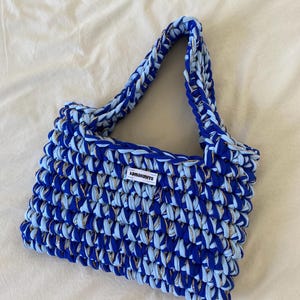 May include: A rectangular crochet handbag crafted from thick, chunky yarn in alternating shades of royal blue and light blue, interwoven with a thin metallic gold thread. The bag features a single handle and a white rectangular label reading ionaknits.