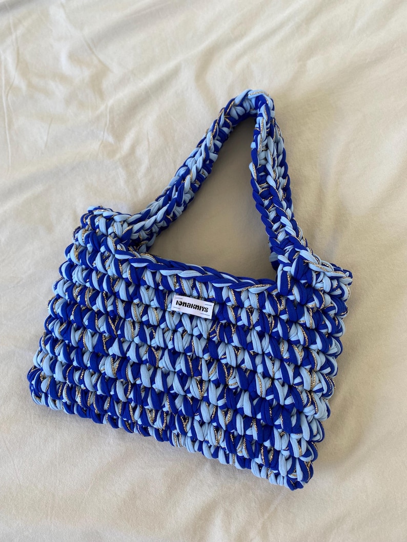 May include: A rectangular crochet handbag crafted from thick, chunky yarn in alternating shades of royal blue and light blue, interwoven with a thin metallic gold thread. The bag features a single handle and a white rectangular label reading ionaknits.