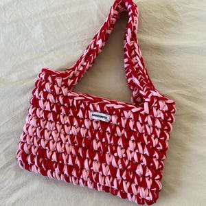 May include: A rectangular crochet handbag crafted from thick, alternating red and light pink yarn. The bag features a single integrated shoulder strap and a small white rectangular fabric label centered on the front with the black text JONAKNITS.