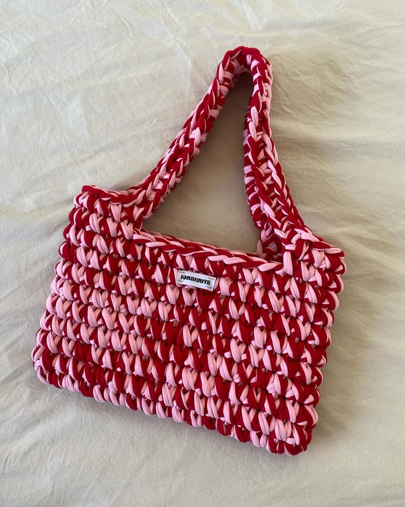 May include: A rectangular crochet handbag crafted from thick, alternating red and light pink yarn. The bag features a single integrated shoulder strap and a small white rectangular fabric label centered on the front with the black text JONAKNITS.
