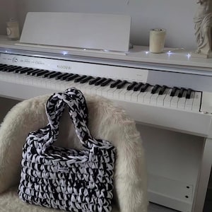 May include: A rectangular handbag crafted from thick, chunky yarn in a marbled black and white pattern. The bag features two braided handles and a small rectangular white label with black text centered on the front exterior surface.