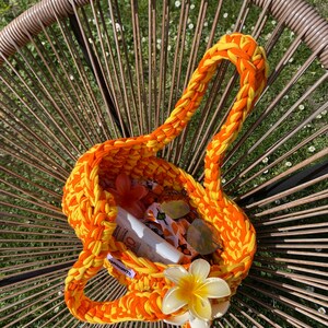 May include: A vibrant orange and yellow crocheted handbag featuring a thick braided handle and a large, cream-colored plumeria flower accent attached to the front. The interior contains a white sunscreen bottle and a patterned fabric pouch.