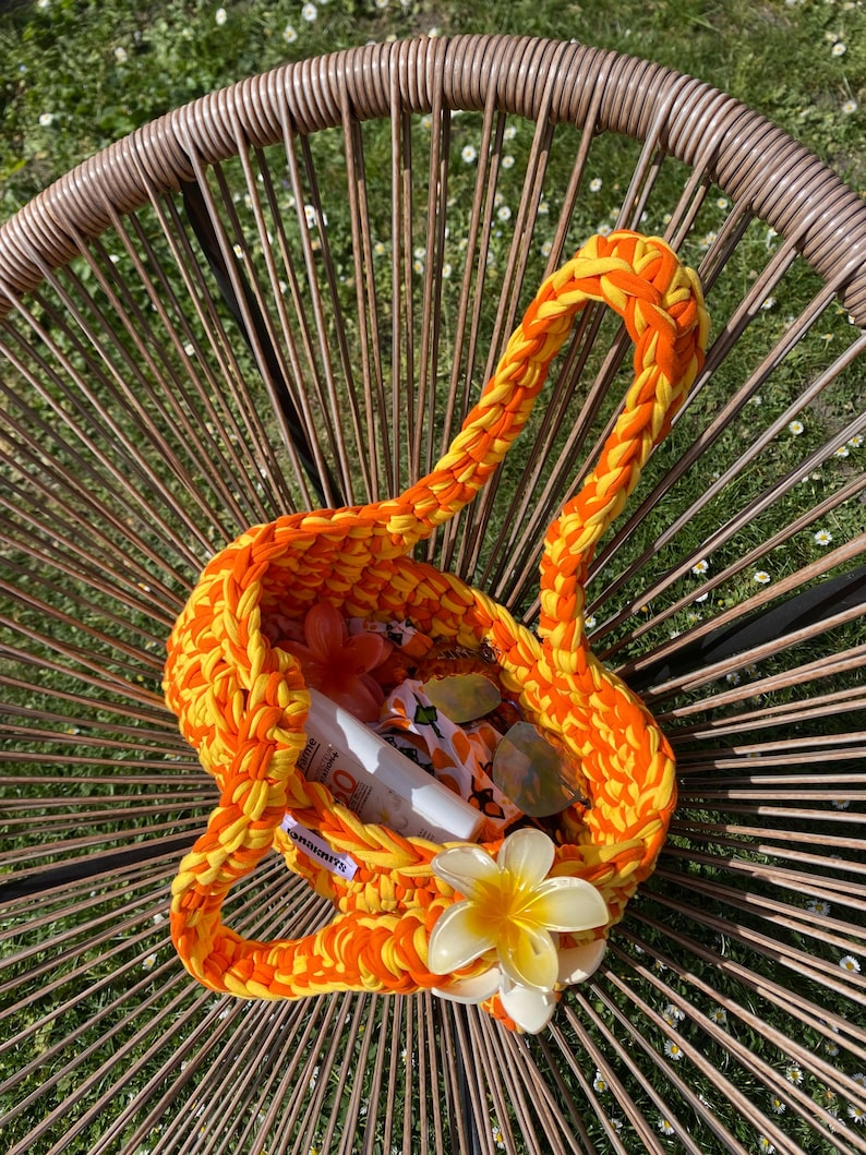 May include: A vibrant orange and yellow crocheted handbag featuring a thick braided handle and a large, cream-colored plumeria flower accent attached to the front. The interior contains a white sunscreen bottle and a patterned fabric pouch.
