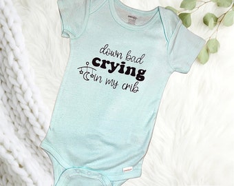 May include: A light blue baby onesie with black text that reads "down bad crying in my crib". A mobile with stars and a crescent moon is above the word "crying".