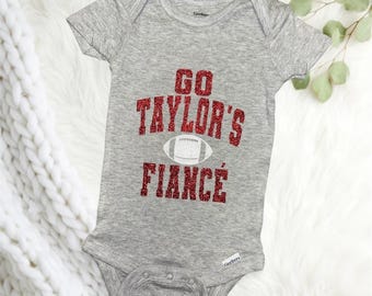 May include: A gray baby bodysuit with short sleeves. The text "GO TAYLOR'S FIANCÉ" is printed in red glitter with a white football graphic in the center. The bodysuit has a snap closure at the bottom.