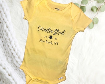 May include: A yellow baby onesie with black text that reads "Cornelia Street 19 89 New York, NY".